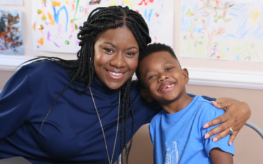 A smiling woman and young boy sit together at a table with a laptop, embraced warmly. Colorful children's drawings decorate the wall behind them.