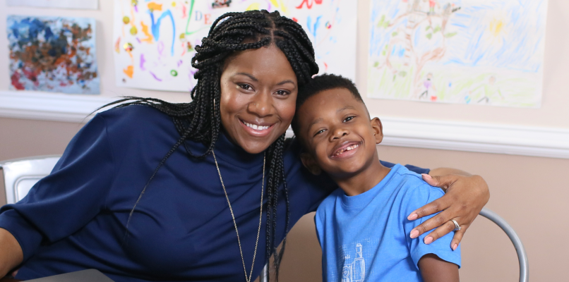 A smiling woman and young boy sit together at a table with a laptop, embraced warmly. Colorful children's drawings decorate the wall behind them.