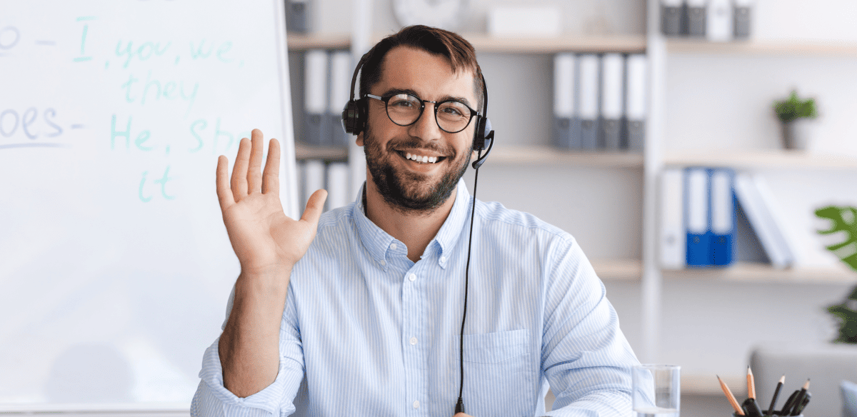 A smiling man in a blue shirt and glasses sits at a desk, raising his hands in a gesture of excitement or celebration.