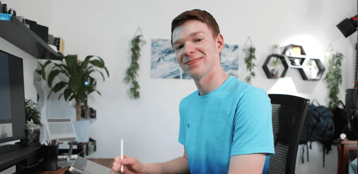 Young man in a blue shirt smiling while holding a stylus in a modern, plant-decorated office. He sits at a desk with a tablet and computer.