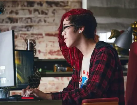 A person with long red hair and glasses is focused at a computer in a cozy room with brick walls. They wear a plaid shirt, conveying concentration.