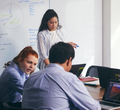 A woman stands by a whiteboard covered in notes, talking to two seated individuals using laptops. The scene conveys cooperation and focus.