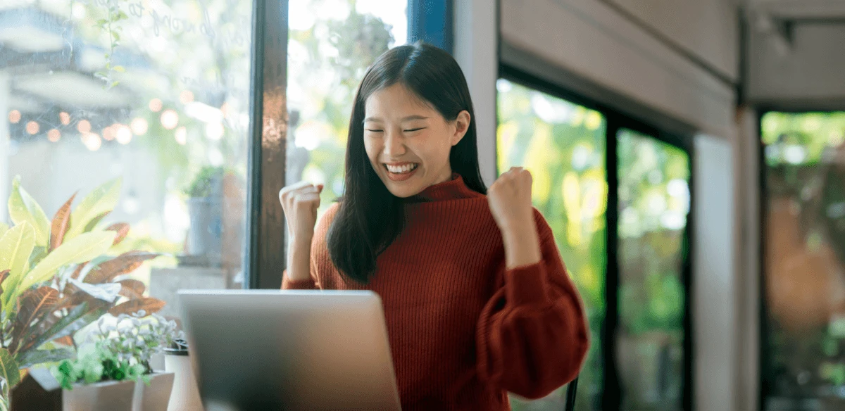 A woman in a cozy red sweater clenches her fists in excitement while looking at a laptop. She is sitting by a window with greenery outside, conveying joy.
