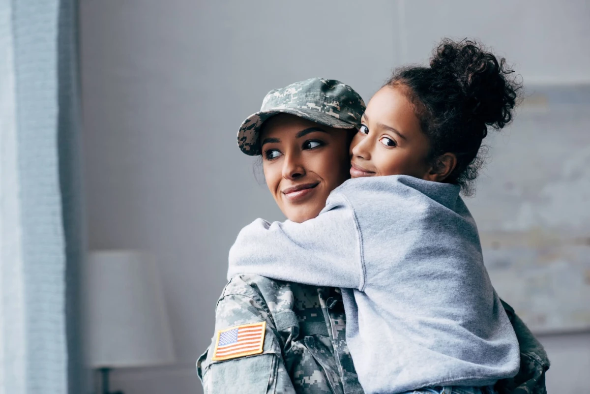 A woman in military uniform with an American flag patch smiles as a young girl hugs her. The scene conveys warmth and affection.