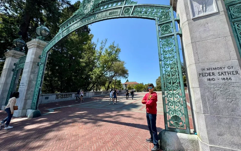 A person standing near the base of a large decorative archway on a university campus. The arch is made of ornate green metal and spans a wide walkway where several people are walking. Tall trees and campus buildings are visible in the background, and a stone pillar beside the arch displays an inscription reading “In Memory of Peder Sather 1810–1886.